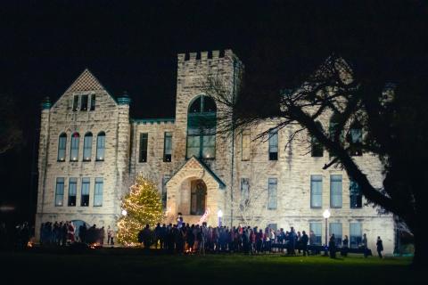 Historic building with people standing in front of it with a large Christmas tree with Christmas lights in front of the building